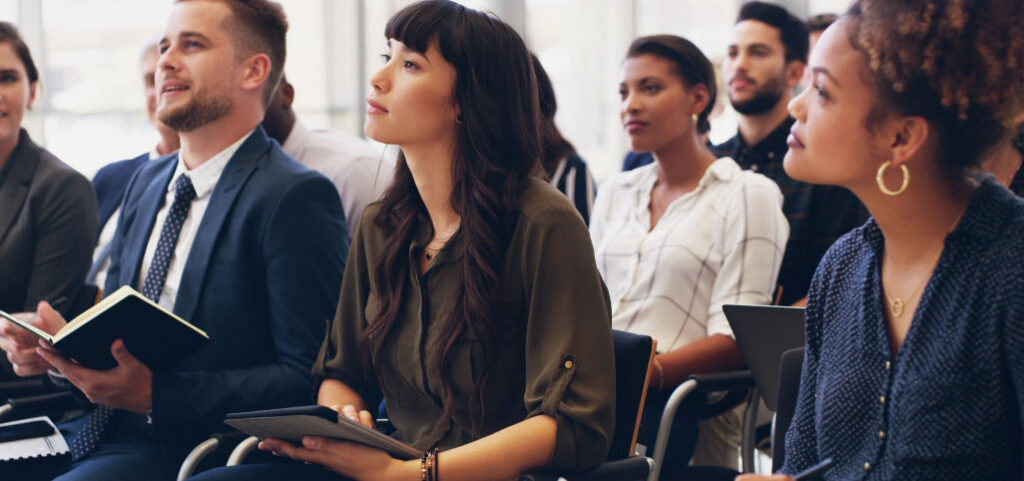 A group of people sits in rows at a seminar or class, attentively listening and taking notes. They appear focused and engaged, fostering a culture of accountability with notebooks and digital tablets in hand.