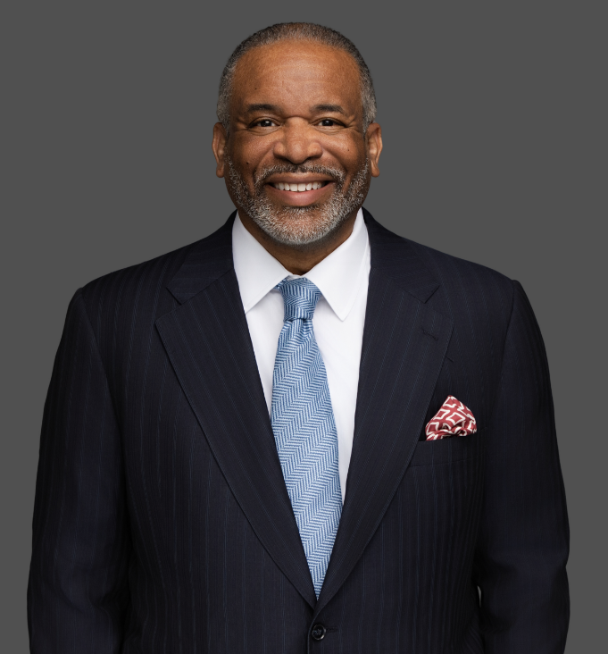 Attorney Gregory J. Vincent with short gray hair and a beard wears a dark pinstripe suit, white shirt, blue patterned tie, and a red-and-white pocket square, standing against a plain gray background.